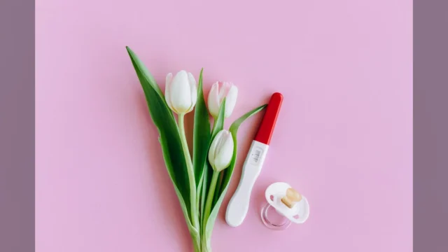 Flat lay of pregnancy test, pacifier, and tulips on pink surface conveying fertility and new beginnings.